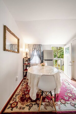 A dining area with a round table, white chairs, a lace tablecloth, a floral rug, a mirror with an ornate frame, a lamp, and a view of the outdoors through a window with curtains.