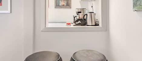 A room with two bar stools, a window with a view of a kitchen, and a framed artwork on the wall.