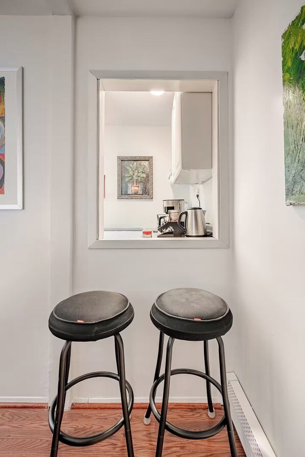 A room with two bar stools, a window with a view of a kitchen, and a framed artwork on the wall.