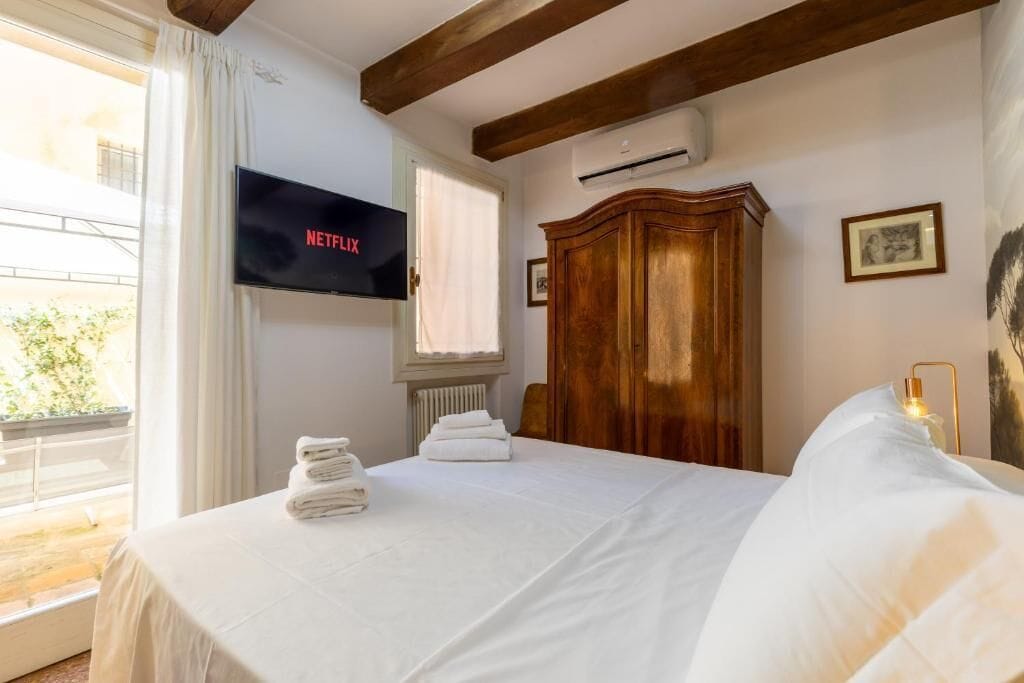Una habitación de hotel con una cama, un televisor mostrando 'NETFLIX', un armario de madera y una ventana con cortinas.