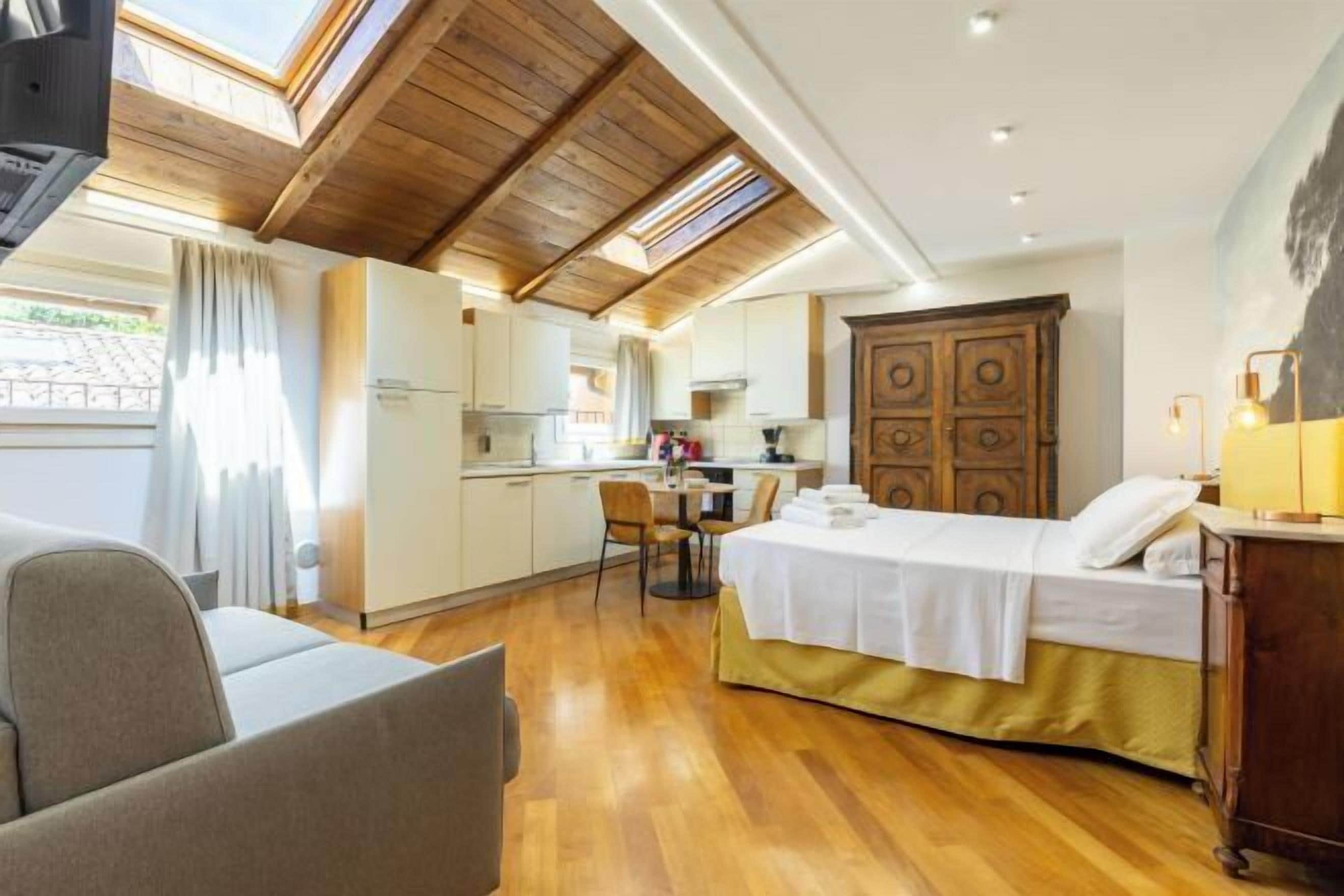 A compact hotel room with a bed, a small kitchenette, a dining area, and a view through a skylight.