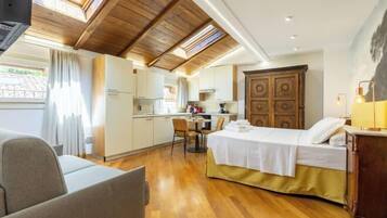 A compact hotel room with a bed, a small kitchenette, a dining area, and a view through a skylight.