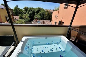 Un jacuzzi con productos de baño azules, una silla de mimbre y vistas a edificios y árboles cercanos.