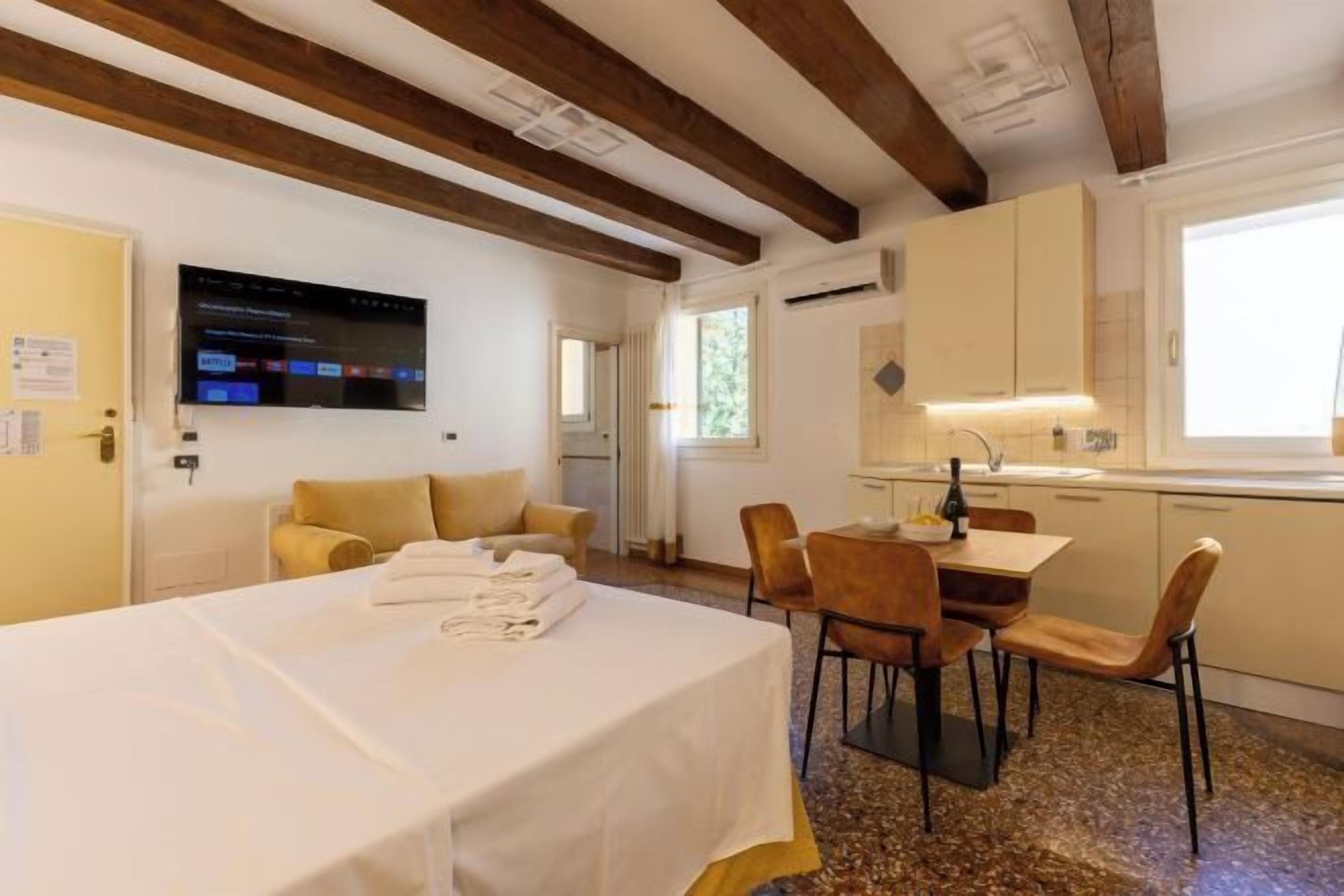 A hotel room with a bed, a dining table with chairs, a TV mounted on the wall, and a kitchen area with cabinets and a window.