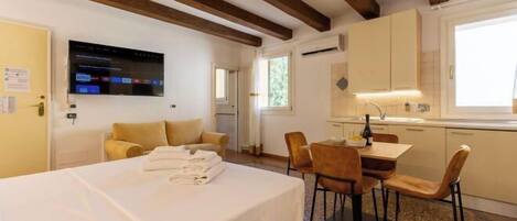 A hotel room with a bed, a dining table with chairs, a TV mounted on the wall, and a kitchen area with cabinets and a window.