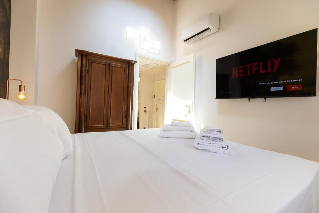 A hotel room with a bed, a TV mounted on the wall displaying 'NETFLIX,' a wooden wardrobe, and a wall-mounted air conditioner.