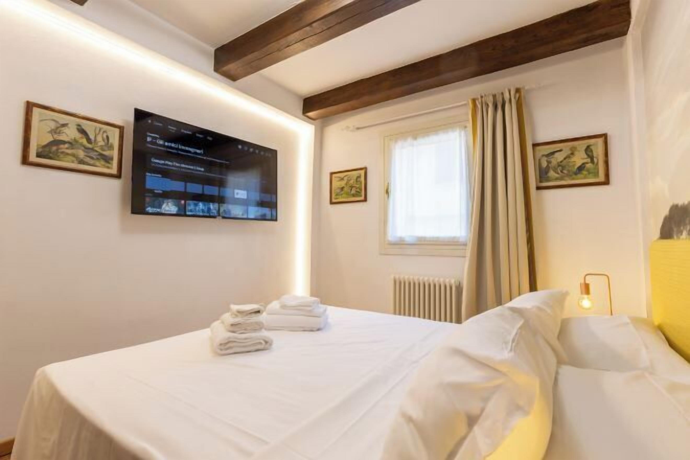 A hotel room with a bed, a flat-screen TV mounted on the wall, and a window with curtains.