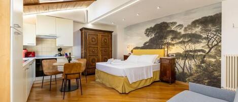 A bedroom with a bed, a sofa, a dining area with chairs, a kitchen, and a mural on the wall.