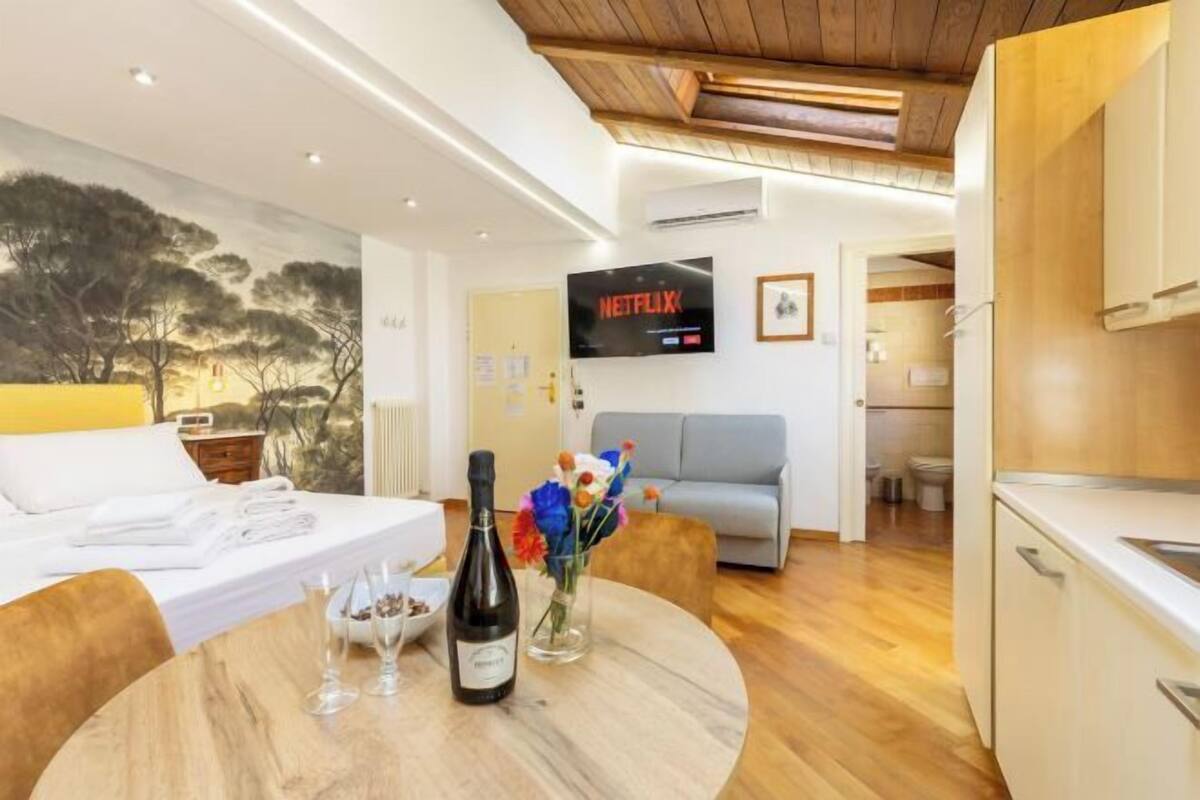 A modern hotel room with a bed, a round dining table with wine glasses and a bottle, a TV displaying Netflix, and a view of the outdoors.