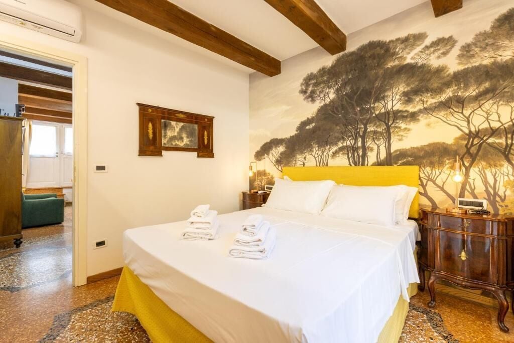 A hotel room with a bed, a nightstand, a wall mural, and a door leading to another room.