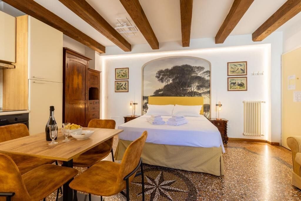 A bedroom with a bed, wooden ceiling beams, a dining table with chairs, and artwork on the walls.