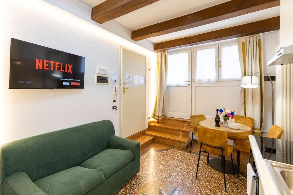 A room with a green sofa, a dining table with chairs, a kitchen area, and a television mounted on the wall displaying 'NETFLIX'.