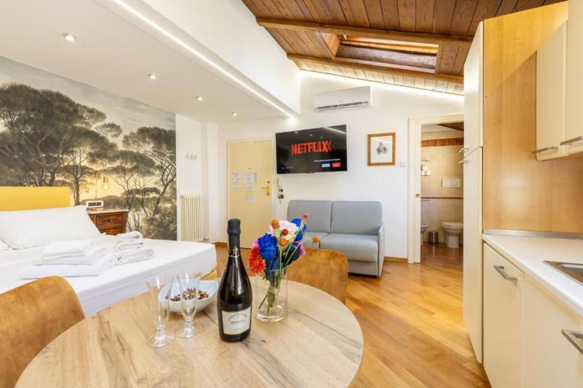 A modern hotel room with a bed, a dining table with wine, a TV, and a view of trees.