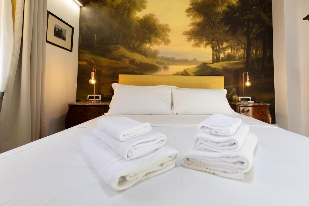 Une chambre d’hôtel avec un lit, du linge de lit blanc, une tête de lit, une table de chevet, une lampe et une fresque murale.