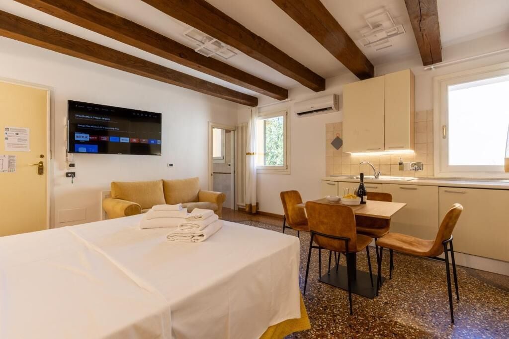 A hotel room with a bed, a dining area with chairs, a TV mounted on the wall, and a kitchen area.