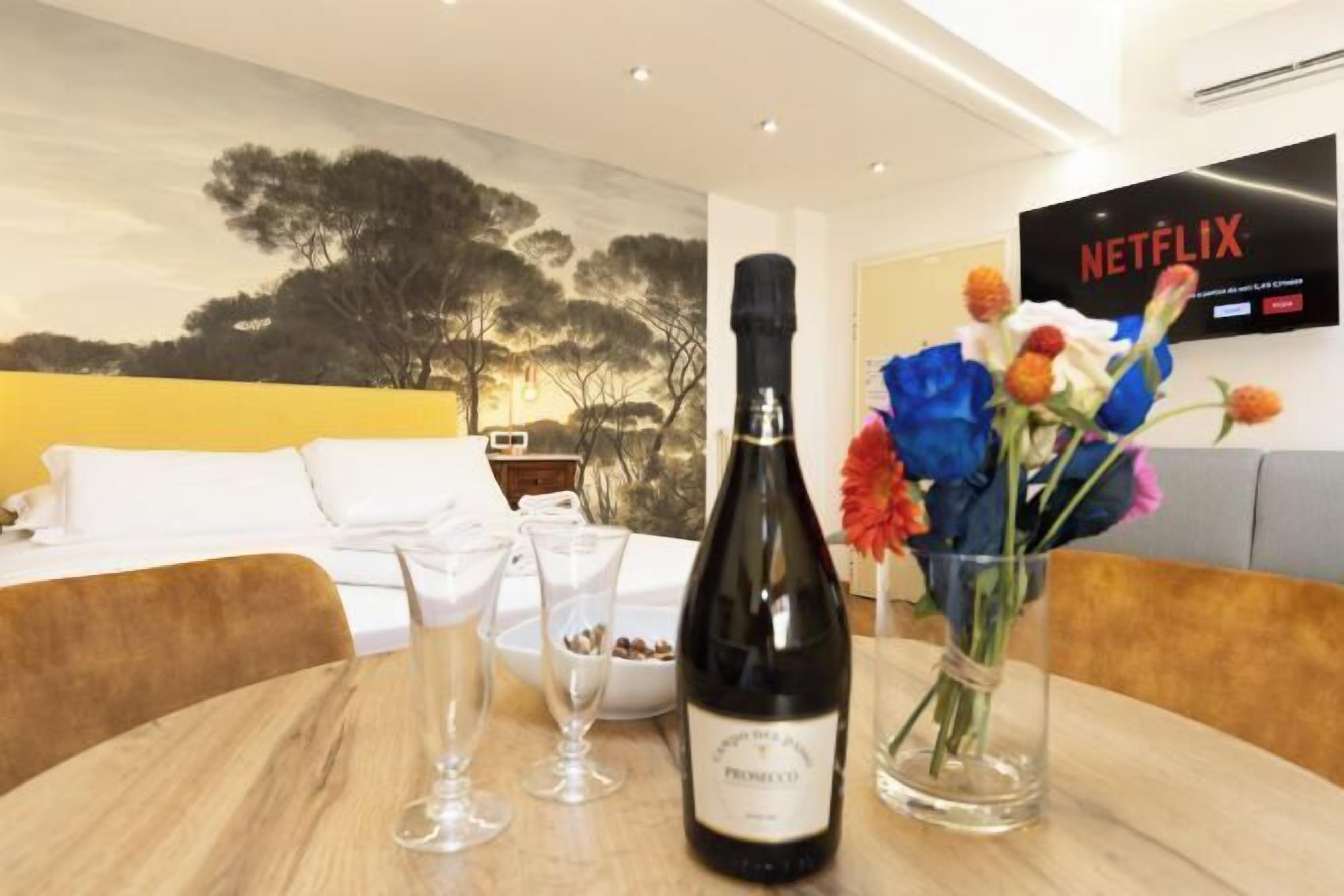 A hotel room with a bed, a table with a bottle of wine, glasses, and a vase of flowers.