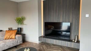 Ein modernes Wohnzimmer mit großem Flachbildfernseher an einer Wand aus Holzpaneelen, einem grauen Sofa mit orangefarbenem Kissen, einem runden Couchtisch mit Dekoration und einer Topfpflanze.