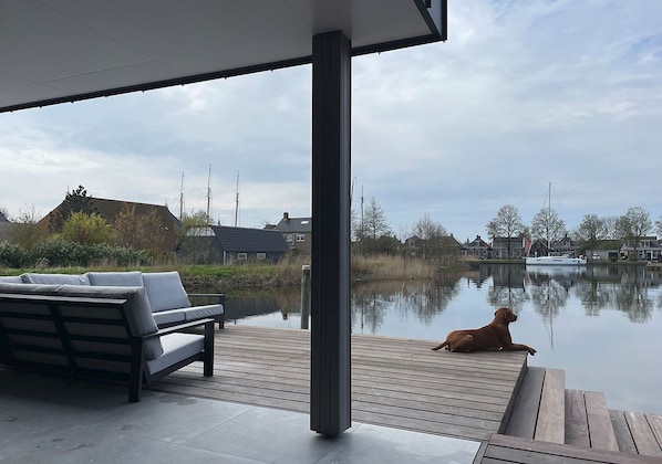 A wooden deck with a bench and a dog, overlooking a calm water body with houses in the background.