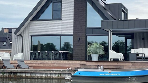 Ein modernes Haus mit großen Fenstern, einem Balkon mit Gartenmöbeln und einem vor der Tür liegenden Boot.