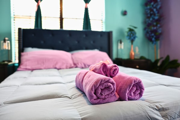 A neatly made bed with pink towels and a dark headboard in a room with teal walls and a window with curtains.