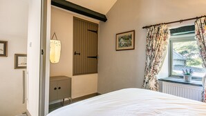 Un dormitorio con cama, armario, mesita de noche pequeña, ventana con cortinas florales y un cuadro en la pared.