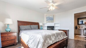 A bedroom with a wooden bed, a ceiling fan, a nightstand with a lamp, a dresser, and a bathroom visible through an open door. - Sunset Getaway on Long Beach (Wainfleet)