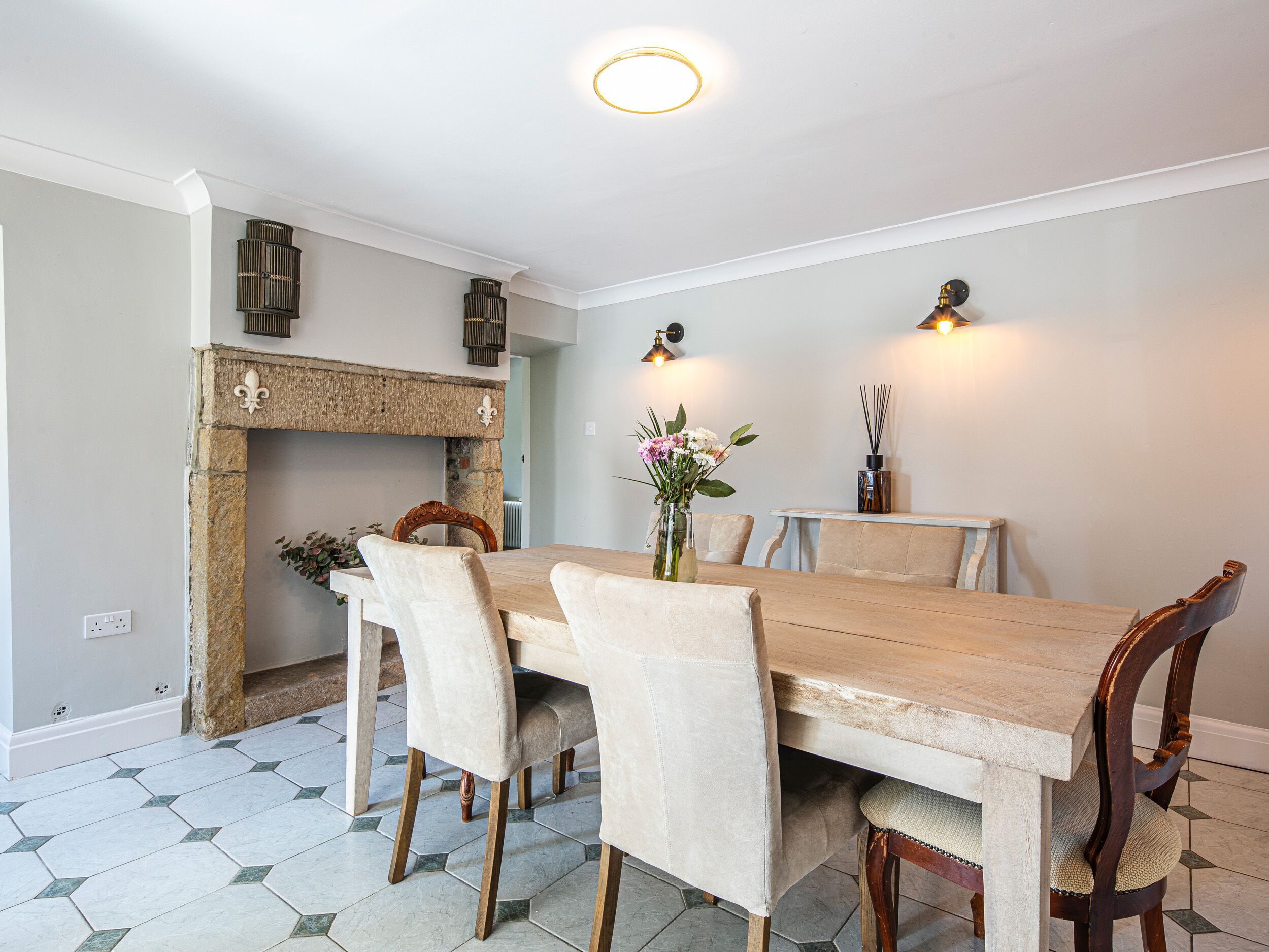 A dining room with a wooden table, beige upholstered chairs, a stone fireplace, and hexagonal patterned floor tiles.