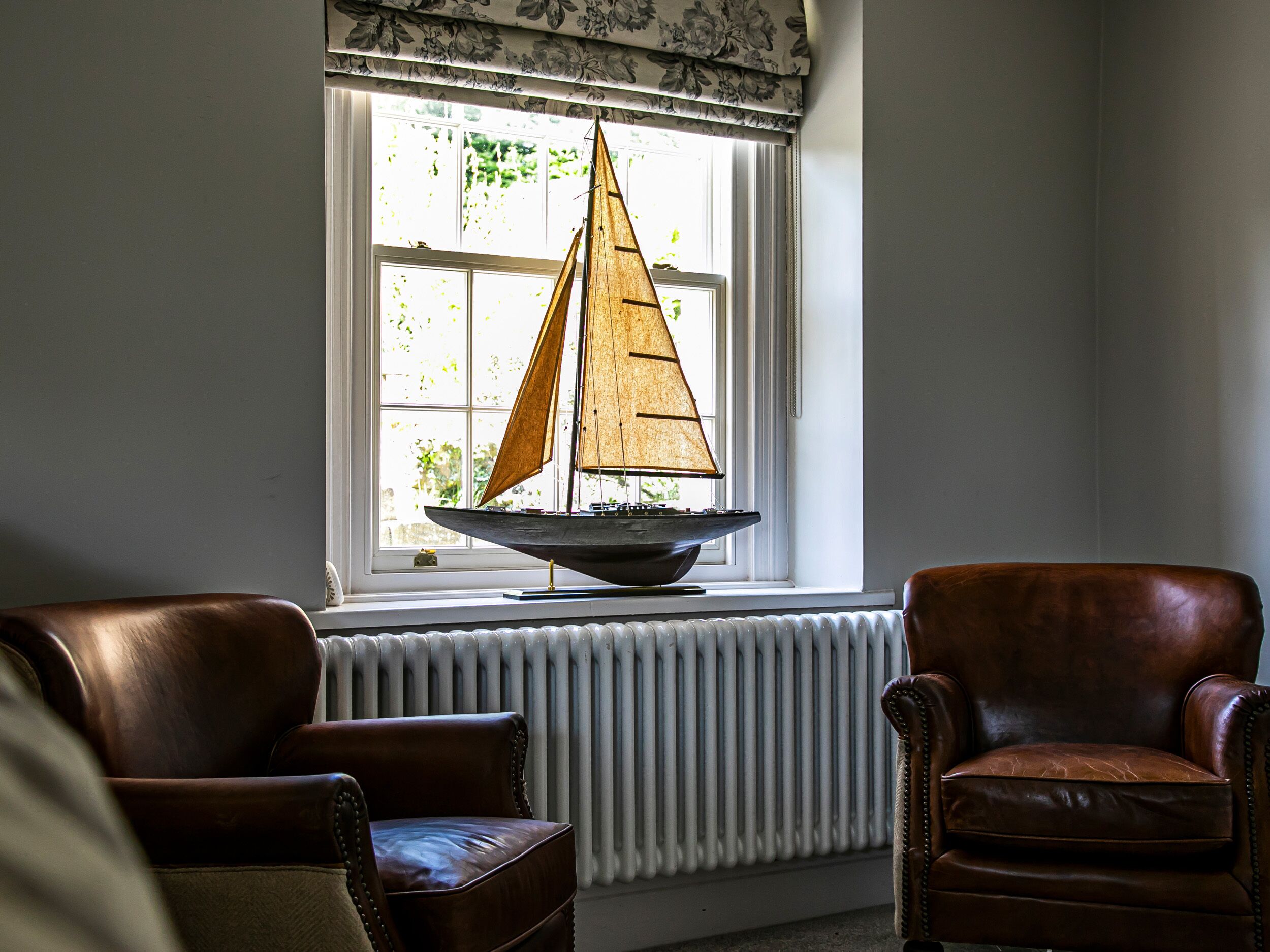 A room with a large window, a model sailboat on a windowsill, and two leather chairs.