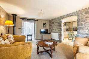 Una sala de estar con un sofá, una mesa de centro y una televisión. Se ve una pared de piedra y una cocina a través de un arco.