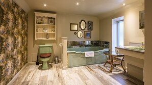 Un baño con inodoro verde, bañera con cerramiento de vidrio, una silla de madera y un tocador con lavamanos.