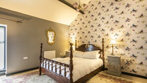 Un dormitorio con una cama de madera con dosel, alfombra estampada con motivos florales, una pared con papel tapiz de pájaros y una mesita de noche con lámpara.