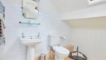 A modern bathroom with a white sink, toilet, and mirror. There is a glass shelf above the sink and a wicker basket on the floor.