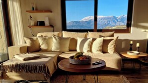 A cozy living room with a beige sectional sofa, a small round coffee table with fruits, a side table with candles, and a large window offering a mountain view.