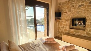 A bedroom with a large bed, a stone wall, a TV mounted on the wall, and a view of a pool outside.