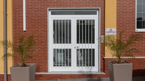 Un bâtiment à la façade en briques rouges, une porte blanche avec des barreaux en métal et deux plantes en pot placées au sol.