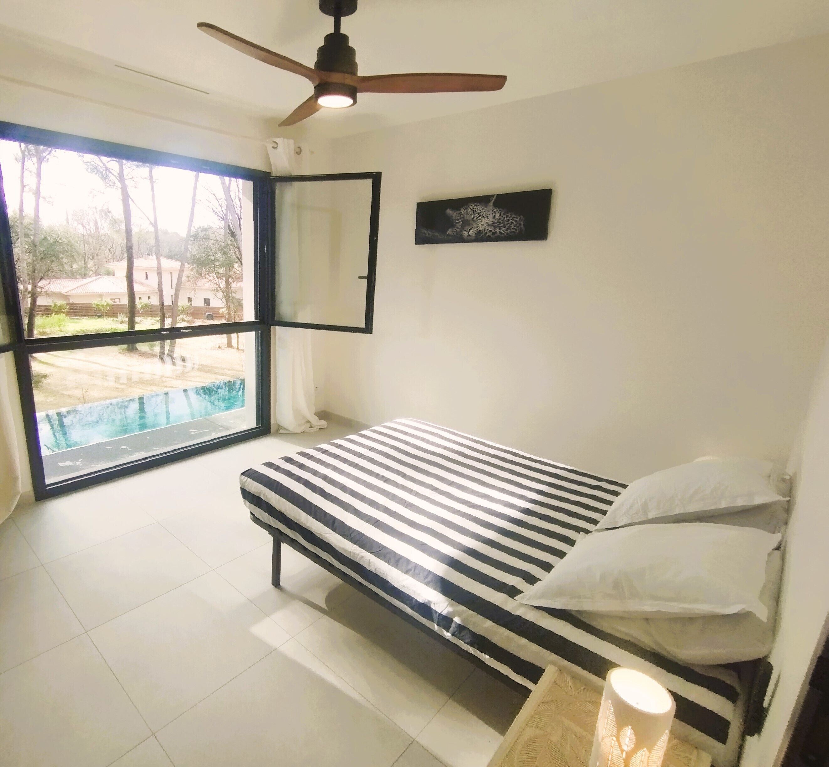 A bedroom with a bed, a ceiling fan, a window with a view of a swimming pool, and a framed picture on the wall.