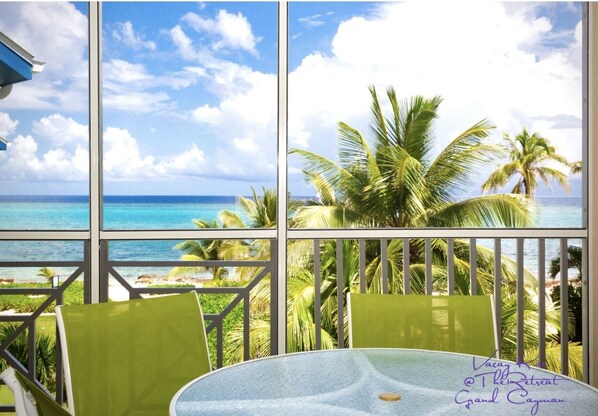 A tropical view from a balcony with a glass table and chairs, overlooking a beach and palm trees.
