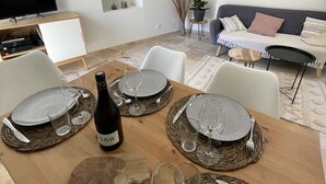 Uma área de jantar com uma mesa de madeira posta para quatro pessoas, rodeada por cadeiras brancas. Há uma garrafa de vinho e copos sobre a mesa.