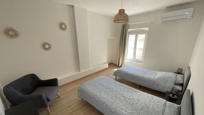 Quarto moderno e minimalista com duas camas de solteiro, uma poltrona cinza e uma pequena mesa com abajur.
