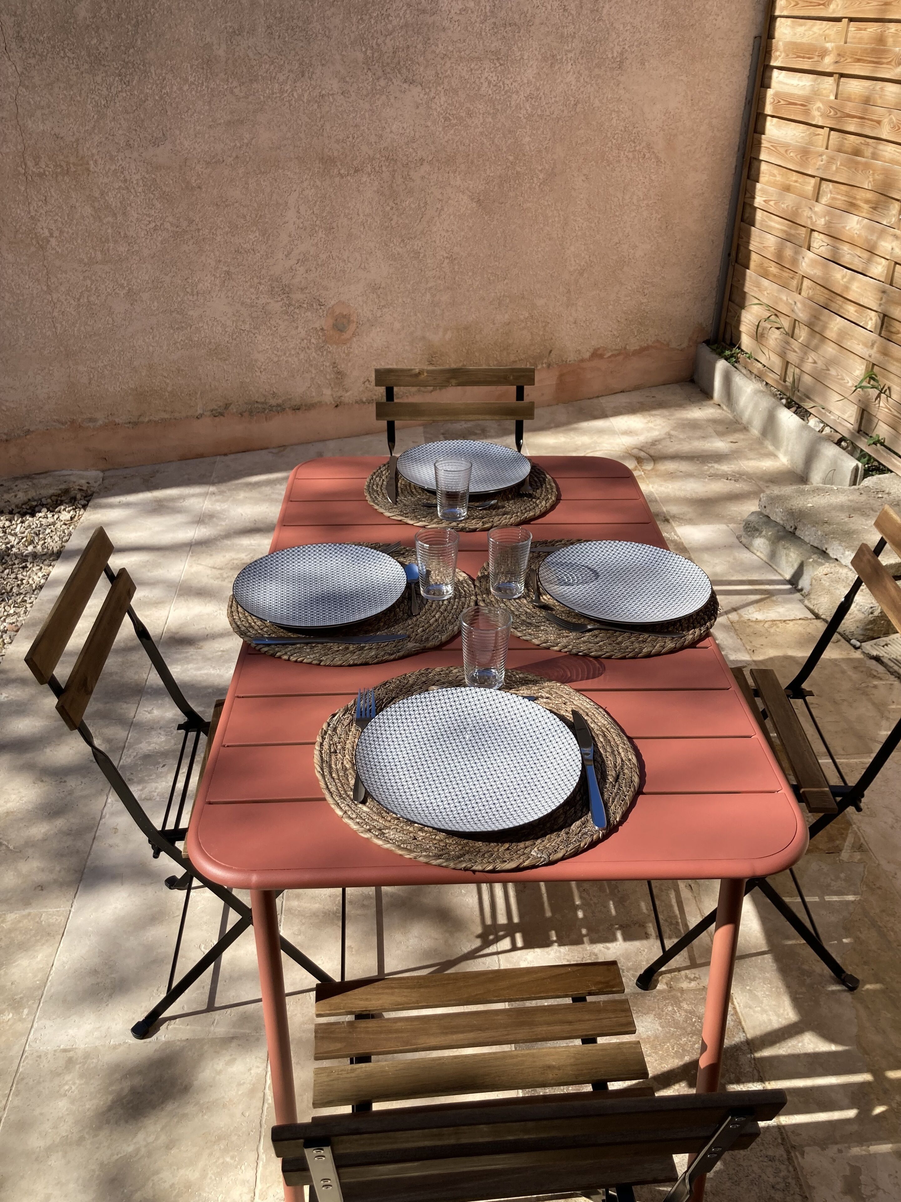Une table en métal rouge, dressée pour quatre personnes, avec des sets de table en fibres tressées, des couverts en métal et des verres transparents.
