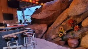 A bar area with stools, a hammock, and a rustic stone wall with potted flowers.