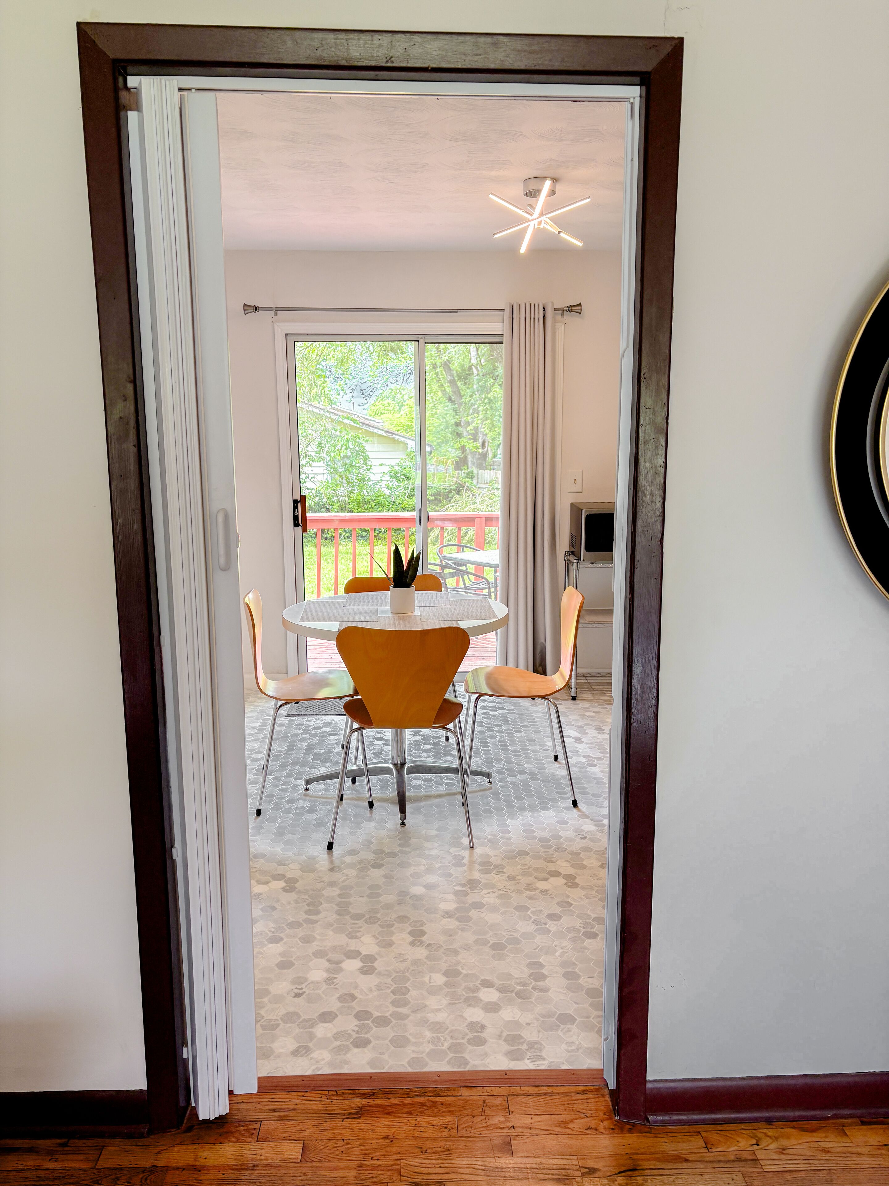 A doorway leading to a dining area with a round table and orange chairs, a ceiling light fixture, and a view of a balcony with a red railing.