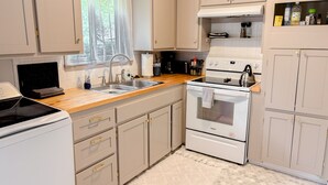 A kitchen with white cabinets, a stove, a sink, and a window with sheer curtains. - Good Vibes House - Centrally located, cozy house!  (Springfield)