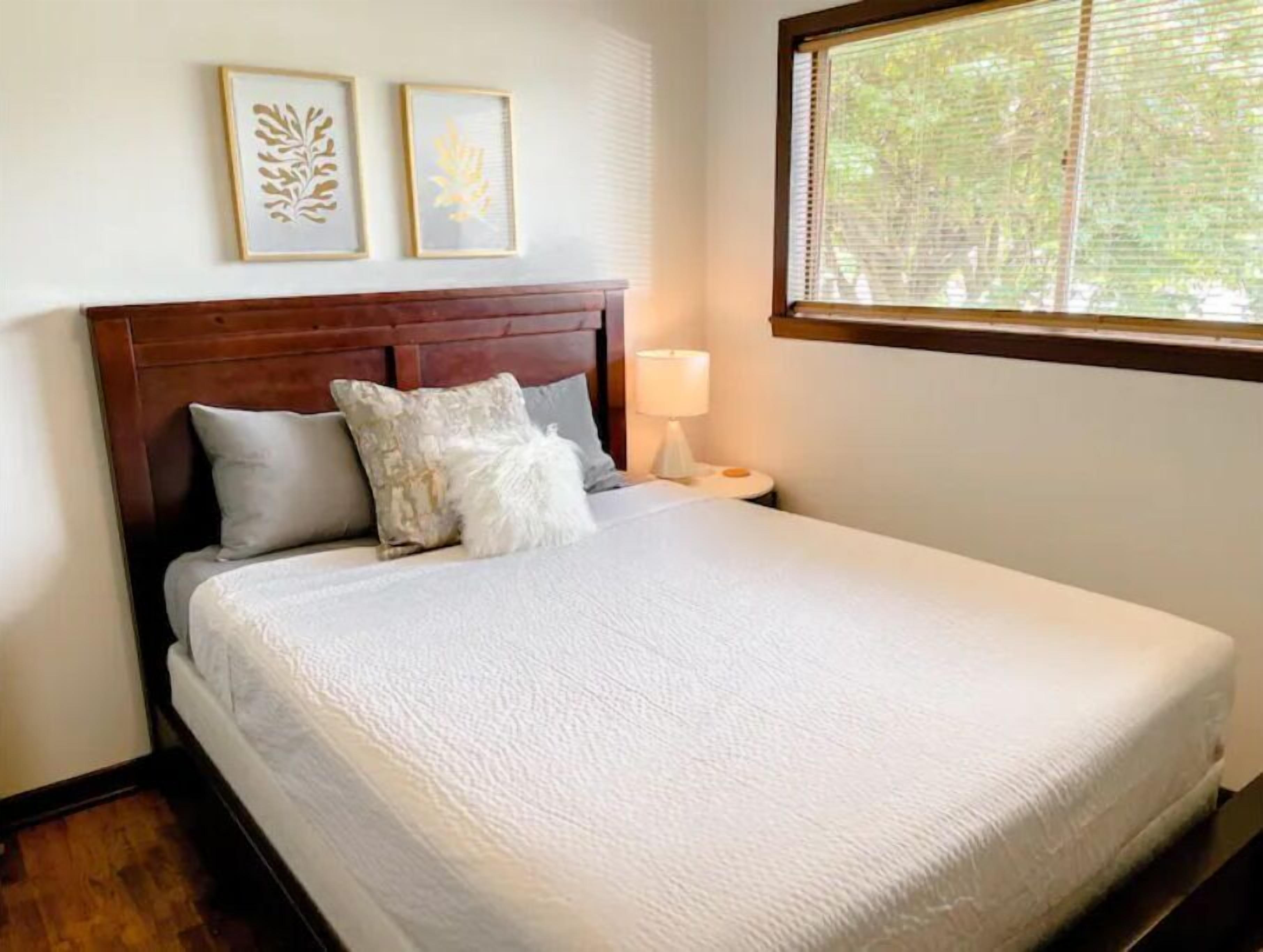 A neatly made bed with a white comforter and pillows, a wooden headboard, and a bedside table with a lamp. There are two framed leaf designs on the wall above the bed.