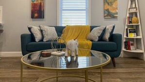 Ein Wohnzimmer mit einem türkisfarbenen Sofa, gemusterten Kissen, einem Glastisch und einem weißen Hängeregal.