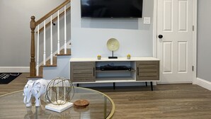 Ein modernes Wohnzimmer mit einem an der Wand befestigten Flachbildfernseher, einer Holztreppe, einem weißen Konsolentisch mit Dekoration und einem runden Glastisch.