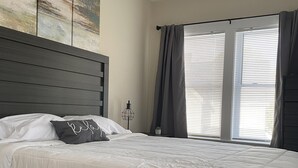 Ein Schlafzimmer mit einem großen Bett, einem Kopfteil, einem Nachttisch, einem Fenster mit Vorhängen und einem Deckenlüfter.