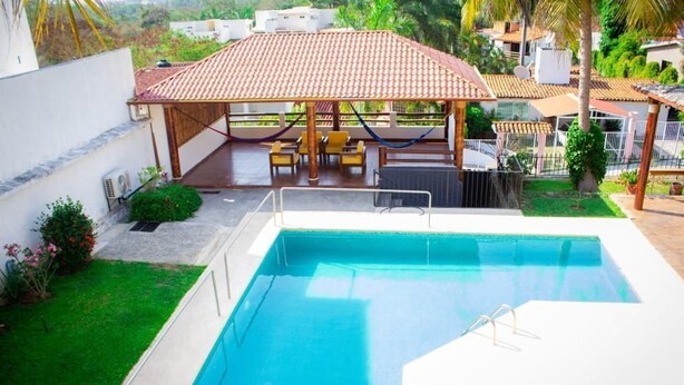 A rectangular swimming pool with a white deck, a covered patio area with yellow chairs, and a view of surrounding greenery and buildings. - Wonderful residence w/studio, 15 min walk distance from beach & restaurants! (Ixtapa Zihuatanejo)