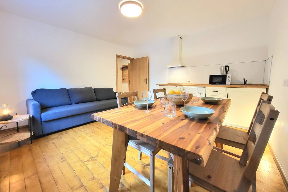 Un espacio habitable pequeño con una mesa de comedor de madera, un sofá azul y una kitchenette al fondo.