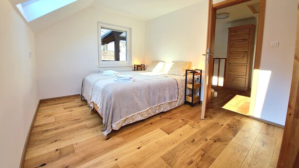 Un dormitorio con tragaluz, piso de madera, cama con ropa de cama, mesita de noche con lámpara y armario de madera.